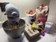 A KitchenAid mixer stands on the left with cookie batter inside. To the right are ingredients for chocolate chip cookies: eggs, vanilla, sugar, flour, brown sugar, chocolate chips, baking powder and baking soda.