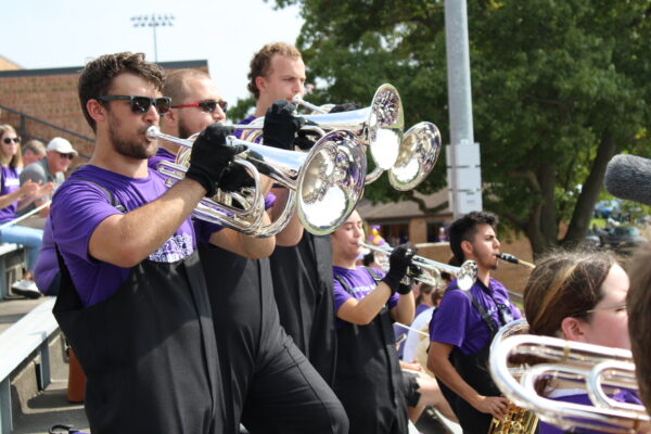 Three young adults wearing black overall pants and purple T-shirts play large silver-brass instruments, facing right. They stand in a staggered row, and more people playing instruments surround them.
