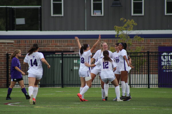 A group of young women in white sports wear stand next to each other on a green turf field, smiling. One has her hands raised in the air, while another rests her hand on her back, leaning towards a smiling player wearing the number 29. Another player wearing purple walks towards the group in the background, sighing.