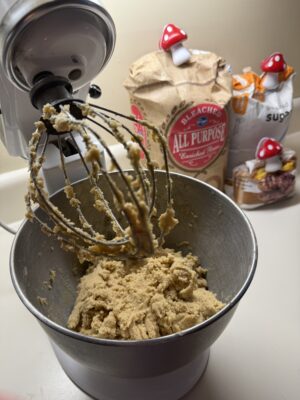 A KitchenAid mixer stands with cookie batter in the mixing bowl. In the background is all purpose flour, sugar and brown sugar.