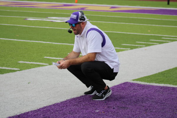 A man with a close-cut beard wearing sunglasses and a baseball cap equipped with a headset crouches on a turf field. He is speaking into the mic, holding a paper and pencil.
