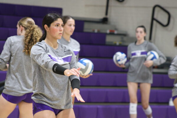 A young woman with long, dark hair pulled back holds her arms out, her expression serious as she looks off to the side of the camera. Behind her are several other young women holding volleyballs in front of purple bleachers, all out of focus.