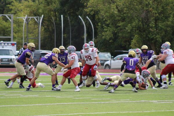 A group of football players in full gear run, caught in motion while one player tackles another in the right of the group. The players are spread out on a green and white turf field with trees in the background, some player’s gear is white and red while others is purple and gold.