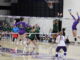 Several women wearing purple and white sports gear are captured mid-jump on a hardwood volleyball court. On the other side of the net, players wearing green move towards a volleyball in the air over the net, where one player in purple leaps, one arm drawn back.