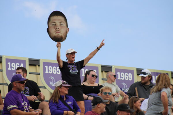 An adult woman smiles while yelling, standing with one hand holding a large sign of a man’s face, the other hand pointing towards the sky. Behind her is a fence with shields showing different years in white against purple, and around her are several rows of people wearing sunglasses or purple and gold clothing, facing forward.