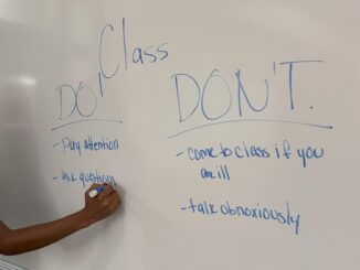 In a classroom, a Black female stands at a large whiteboard, writing with a blue Expo marker. On the whiteboard reads ‘Class Do and Don’t’ followed by a list of behaviors and actions that should and should not be seen in the classroom.