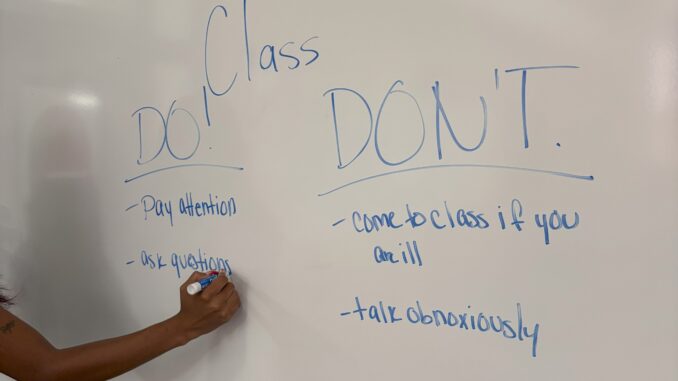 ClassroomWhiteboard(1) In a classroom, a Black female stands at a large whiteboard, writing with a blue Expo marker. On the whiteboard reads ‘Class Do and Don’t’ followed by a list of behaviors and actions that should and should not be seen in the classroom.