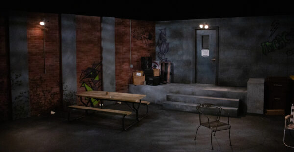 A stage set up to look like the back area of a coffee shop. There are red brick walls with stone slats separating the brick, with various graffiti texts. The lighting is dark and dim, and there is an empty wooden picnic bench, a stray metal chair, and various empty brown boxes.