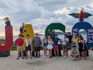 A group of people stand and smile at the camera in front of large colored letters spelling “JACO” with “Costa Rica” written on the “O.” Sculpted animals pose on the letters, including a toucan, a monkey, a sloth, and a red macaw. Behind the group is a cloudy sky over the ocean.