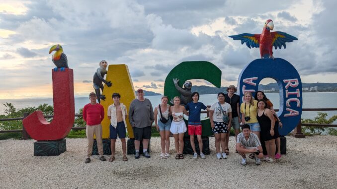 A group of people stand and smile at the camera in front of large colored letters spelling “JACO” with “Costa Rica” written on the “O.” Sculpted animals pose on the letters, including a toucan, a monkey, a sloth, and a red macaw. Behind the group is a cloudy sky over the ocean.