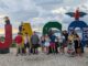 A group of people stand and smile at the camera in front of large colored letters spelling “JACO” with “Costa Rica” written on the “O.” Sculpted animals pose on the letters, including a toucan, a monkey, a sloth, and a red macaw. Behind the group is a cloudy sky over the ocean.