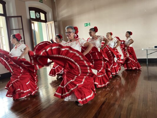 A group of about seven women dressed in white blouses and large, red skirts dance in a room with white walls, a dark hardwood floor, and dark wood trimming the windows. The women are twirling their skirts, and the fabric is slightly blurred as it moves.