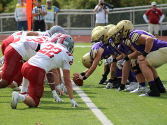 Two lines of football players in full gear crouch facing each other on either side of a yardline, one player holding a football out against the green turf between them. The players on the left are dressed in white and red, while the players on the right are purple and gold.