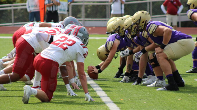 Two lines of football players in full gear crouch facing each other on either side of a yardline, one player holding a football out against the green turf between them. The players on the left are dressed in white and red, while the players on the right are purple and gold.