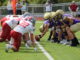 Two lines of football players in full gear crouch facing each other on either side of a yardline, one player holding a football out against the green turf between them. The players on the left are dressed in white and red, while the players on the right are purple and gold.