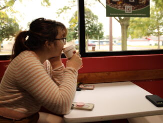 A young adult sits at a white table drinking from a white paper cup. They are wearing a pink and white striped shirt and looking out the window towards the street and nearby trees.