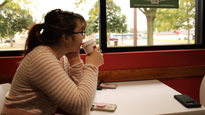 A young adult sits at a white table drinking from a white paper cup. They are wearing a pink and white striped shirt and looking out the window towards the street and nearby trees.