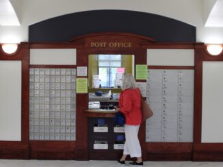 The exterior of a post office. The space features a dark wood facade with the words, “POST OFFICE” displayed above a central window. On either side of the window, there are rows of metallic postal boxes integrated into the wood paneling. An older woman wearing a red shirt and white pants stands at the outside of the window, looking in.
