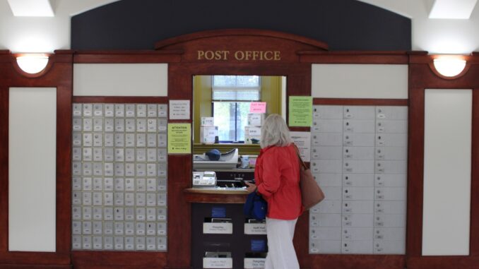 The exterior of a post office. The space features a dark wood facade with the words, “POST OFFICE” displayed above a central window. On either side of the window, there are rows of metallic postal boxes integrated into the wood paneling. An older woman wearing a red shirt and white pants stands at the outside of the window, looking in.