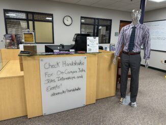 A whiteboard attached to a wooden desk reads, “Check Handshake for On-Campus Jobs, Jobs, Internships, and Events! albion.edu/handshake.” A mannequin wearing gray slacks, a purple-and-white-striped shirt and a gray tie stands to the right of the desk.