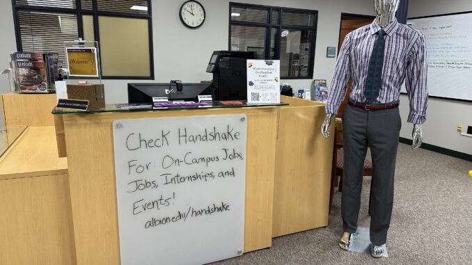 Handshake + mannequin A whiteboard attached to a wooden desk reads, “Check Handshake for On-Campus Jobs, Jobs, Internships, and Events! albion.edu/handshake.” A mannequin wearing gray slacks, a purple-and-white-striped shirt and a gray tie stands to the right of the desk.