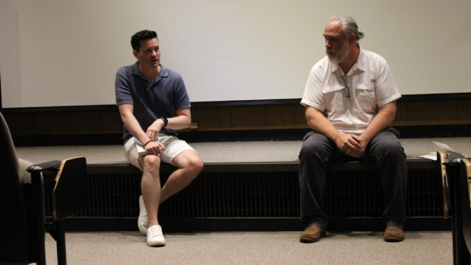 ACReading Two men sit on the edge of a low, carpeted stage, with a blank white screen behind them. The man on the left wears a dark blue short-sleeved button up, and is speaking and holding a book in his lap. The man on the right has a white beard and wears a white short-sleeved button up, and is looking to the right.