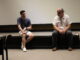 Two men sit on the edge of a low, carpeted stage, with a blank white screen behind them. The man on the left wears a dark blue short-sleeved button up, and is speaking and holding a book in his lap. The man on the right has a white beard and wears a white short-sleeved button up, and is looking to the right.