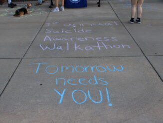 A sidewalk with pastel chalk writing promoting a suicide awareness walkathon reading “1st annual Suicide Awareness Walkathon” and “Tomorrow needs YOU!” Also visible on the pavement nearby are a few pairs of feet where people are standing, as well as a dog lying on the sidewalk.