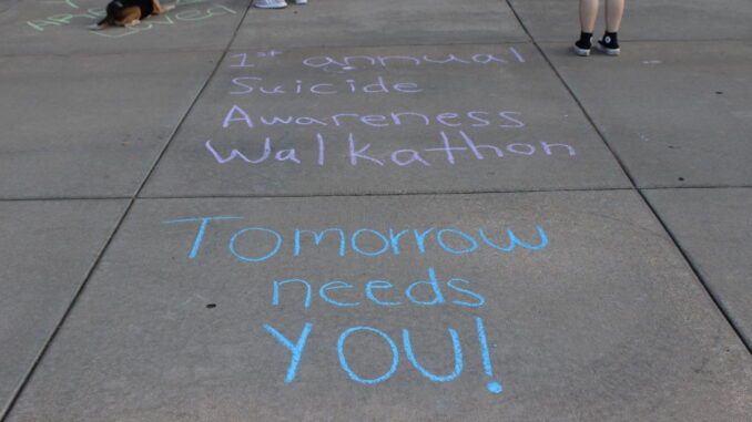 A sidewalk with pastel chalk writing promoting a suicide awareness walkathon reading “1st annual Suicide Awareness Walkathon” and “Tomorrow needs YOU!” Also visible on the pavement nearby are a few pairs of feet where people are standing, as well as a dog lying on the sidewalk.