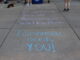 A sidewalk with pastel chalk writing promoting a suicide awareness walkathon reading “1st annual Suicide Awareness Walkathon” and “Tomorrow needs YOU!” Also visible on the pavement nearby are a few pairs of feet where people are standing, as well as a dog lying on the sidewalk.
