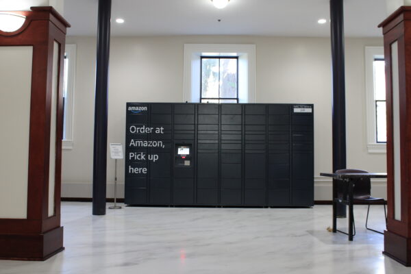 A black set of connected lockers reading “amazon” and “Order at Amazon, Pick up here” sit in front of a set of white walls and windows. The floor underneath the lockers is white marble, with a singular unidentified sign on a metal pedestal placed on it. 