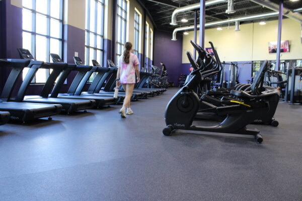 Five black treadmills are displayed in a room with floor to wall windows. The floor underneath the treadmills is purple with white speckles.