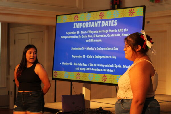 Two young people stand on either side of a large TV screen.  They are both in casual dress; the person farther from the camera is looking at the screen and speaking, while the person nearest to the camera is standing to the side and looking forward. The TV screen displays a blue background with a yellow border, and capitalized white letters near the top of the screen read “important dates.” In the background a low stage, white walls and white doors are visible.