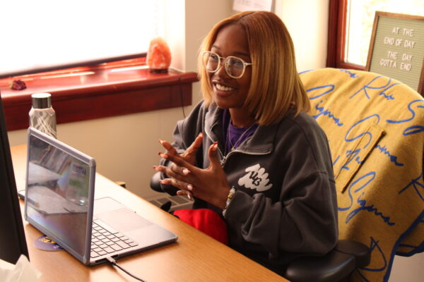A woman sits at a desk with a laptop and a water bottle, smiling and gesturing with her hands. 