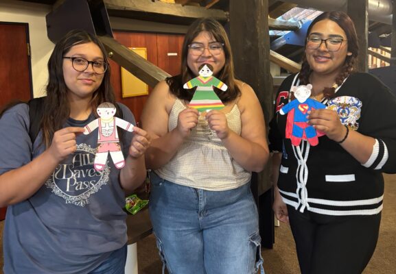 Three young adults stand in a row facing the camera and smiling. They are all wearing glasses and casual clothes, and hold up colorfully decorated flat paper dolls.