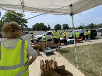 A person in the foreground wearing a bright yellow vest watches ten other people dressed similarly standing at the edge of a parking lot. The people at the curb lift boxes and brown paper bags into the back of a white car parked at the edge.