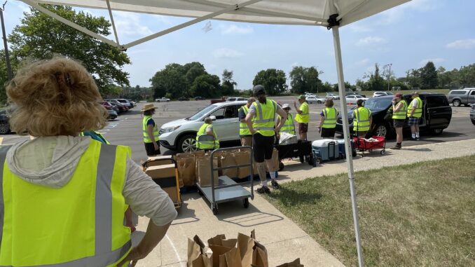 A person in the foreground wearing a bright yellow vest watches ten other people dressed similarly standing at the edge of a parking lot. The people at the curb lift boxes and brown paper bags into the back of a white car parked at the edge.
