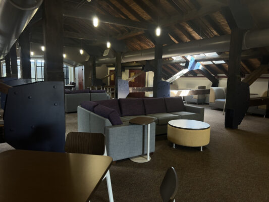 A loft-style space with gray and white sectional sofas, tables, wooden beams and ambient lighting.