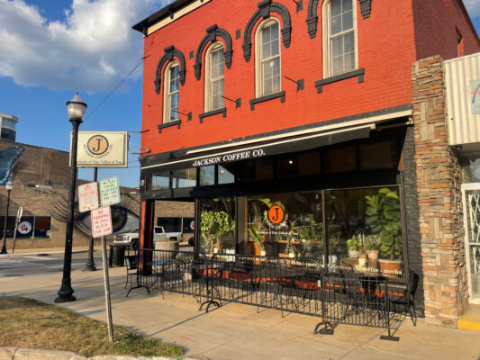 A wide shot of a two-story bright red brick building, with the lower level consisting of three large square windows lined with black edges. The middle window has an orange circle with a black J in the middle and “Jackson Coffee company” written in white under it. Outside next to the windows are four sets of black metal chairs and tables. A matching metal fence separates the tables from the public sidewalk. A no parking sign sits in front of an out of focus Jackson Coffee Company Business sign. 