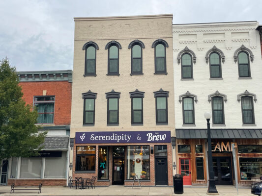 A wide shot of a three-story beige brick building in the center. The lowest level of the building is built in its original red brick. There are two black-lined windows on either side of a black metal door on this level, with a window with a cyan blue bow on the glass. A purple banner sits above the windows with a grape decal  “Serendipity and the Brew” printed on it. In front of the building is a black metal table set right outside, and there are two other buildings sandwiching it in. One is red brick and two stories, the other is white brick and three stories. 