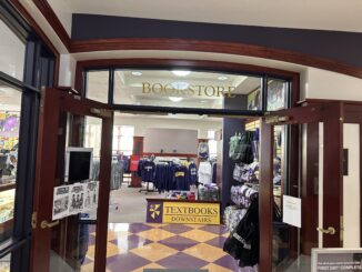 Racks of T-shirts and sweatshirts in a room, with stuffed bears and blankets on the right side of the room. Above the top of the entrance, golden text reads, “Bookstore” on a glass pane.