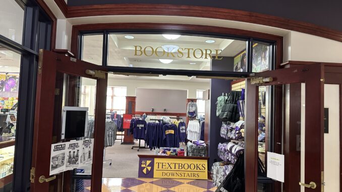 Bookstore Racks of T-shirts and sweatshirts in a room, with stuffed bears and blankets on the right side of the room. Above the top of the entrance, golden text reads, “Bookstore” on a glass pane.