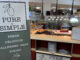 A white and muted green banner stands in the front of a countertop with the words, “Pure and simple” and “Fresh, Delicious, Allergen-free Dining” printed on it. In the background is a stack of plates on a lower shelf and placed above it on the counter are roasted Yukon potatoes, garlic green beans and Montreal Roasted Eye Round. Out of focus, there is a paper displaying the menu and a small kitchen area.