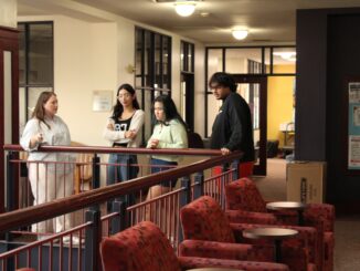A group of three young people look over a railing as a fourth person points. Behind them is an open door surrounded by windows.