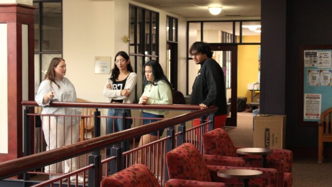 A group of three young people look over a railing as a fourth person points. Behind them is an open door surrounded by windows.