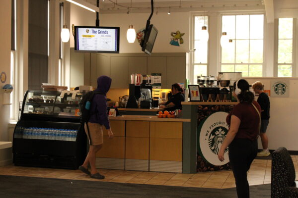 Students walk in front of a storefront with a counter and pastry shelf. On the front of the counter is the Starbucks logo in dark green and white. 