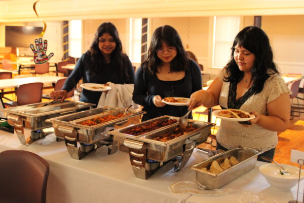 Three young adults with long hair in casual dress face the camera, looking down at a white table in front of them and serving themselves from the silver trays of food in a large, open, sunlit space.