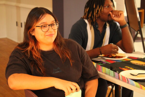 A young person with long hair and glasses sits in a chair beside a table, looking ahead of them, away from the camera, and lightly smiling. Behind them a man is visible sitting at the table eating.