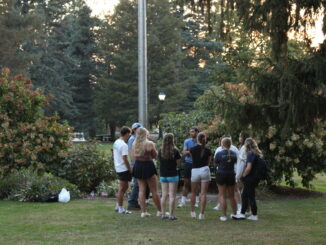 A group of students stands in a circle on a grassy lawn. In front of them are bushes with light pink flowers and a steel flagpole.