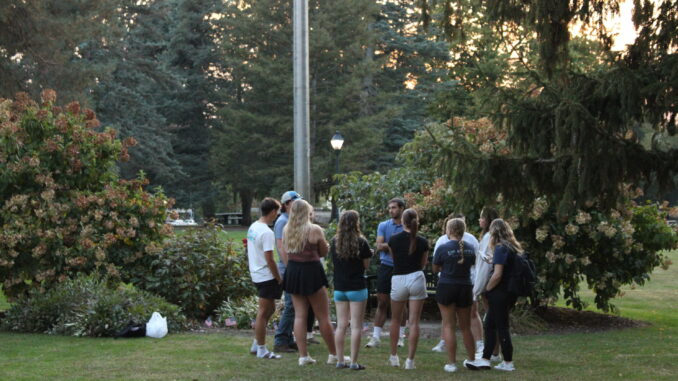 A group of students stands in a circle on a grassy lawn. In front of them are bushes with light pink flowers and a steel flagpole.